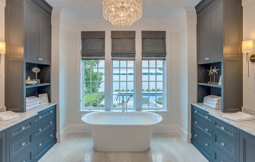 Vanity area with blue and brown cabinets on each side of freestanding bathtub three tall windows and ornate chandelier above