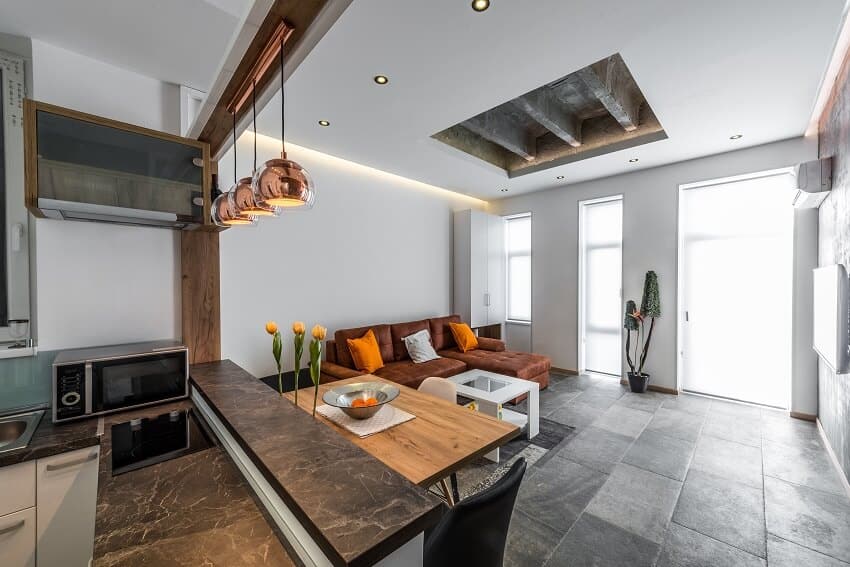 View from kitchen to living room with grey tile floors sofa wood dining table marble kitchen counters and pendant lights in a modern flat interior