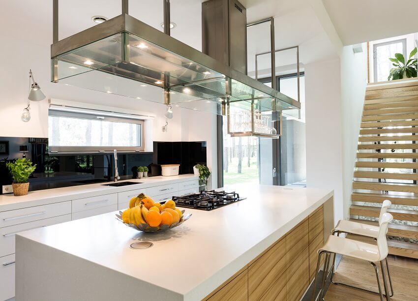 White modern kitchen in trendy house with cooktop on island, and stainless and glass range hood