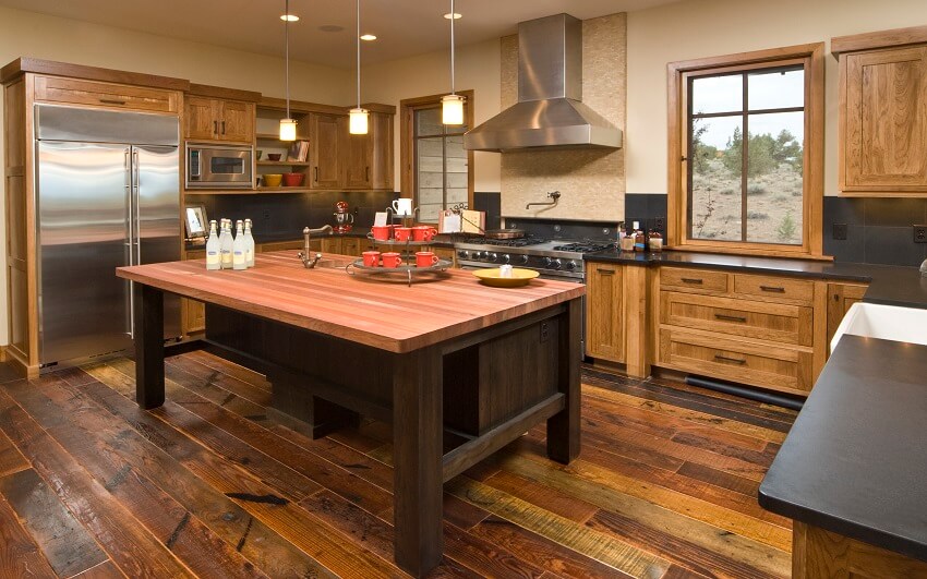 Wood cabinets black countertops black tile backsplash pendant lights stainless steel appliance hardwood floors and island with beech countertop in a rustic kitchen