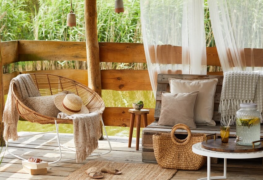 Wooden gazebo by the lake with rattan armchair sheer curtains and a bench with throw pillow and blanket