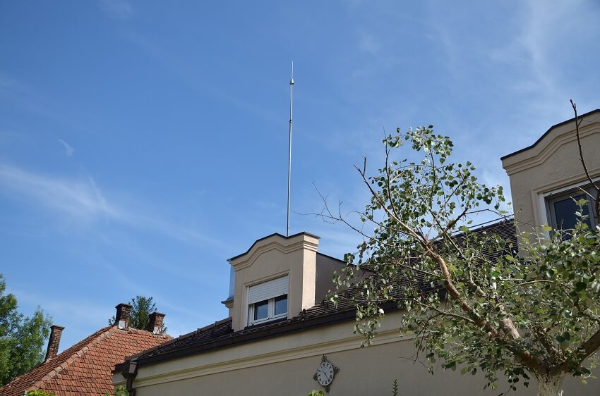 A lightning rod on the roof of a house with a hanging clock on its exterior wall and a tree
