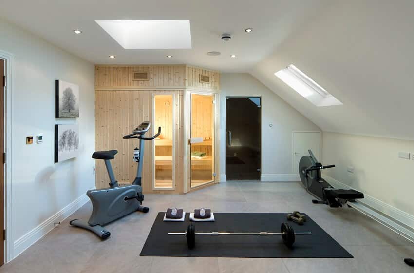 A sauna room a shower cubicle and a home gym with exercise bike rowing machine and some free weights and natural light is streaming in through two skylight windows