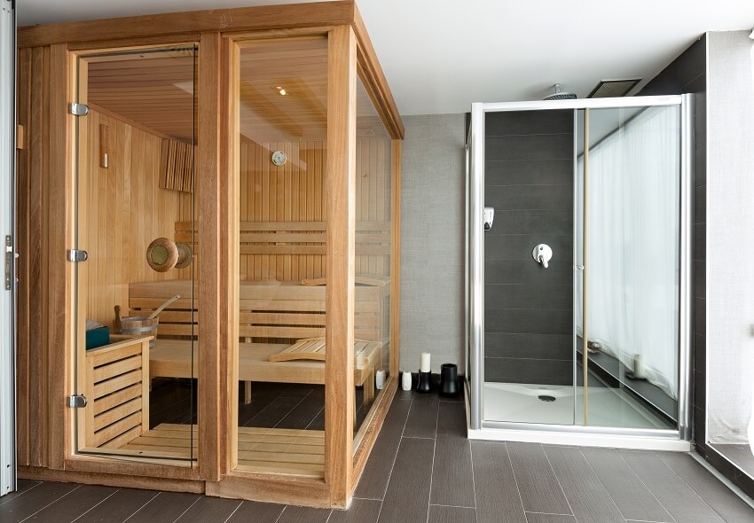 A vinyl floored room with wooden framed sauna cube with wood interiors and benches and a shower area with ceiling mounted shower head