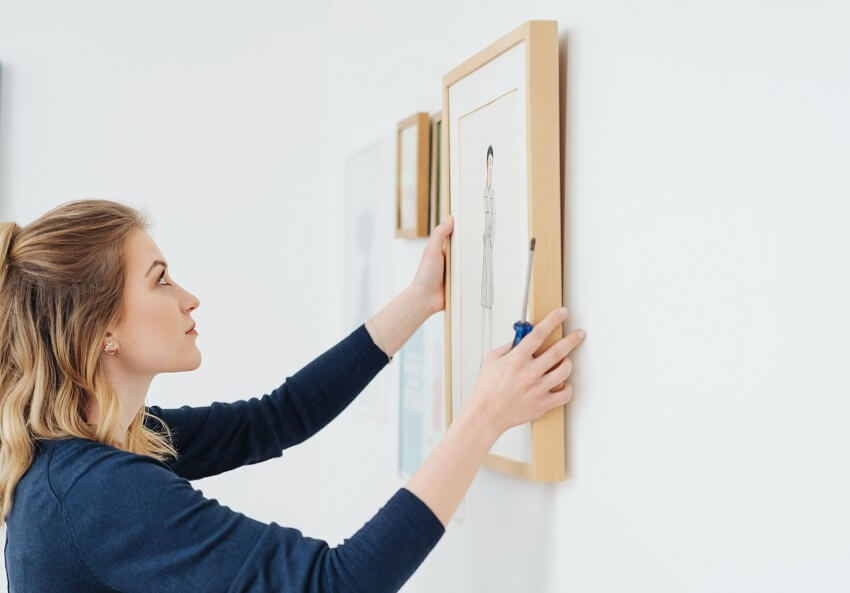 A woman hanging a picture in a plain wooden frame on a white wall