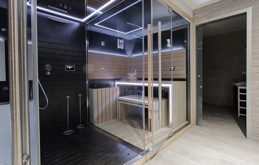 An open shower with ceiling mount rain shower head and a modern home finnish sauna room with neon lights and wood bench enclosed in a glass