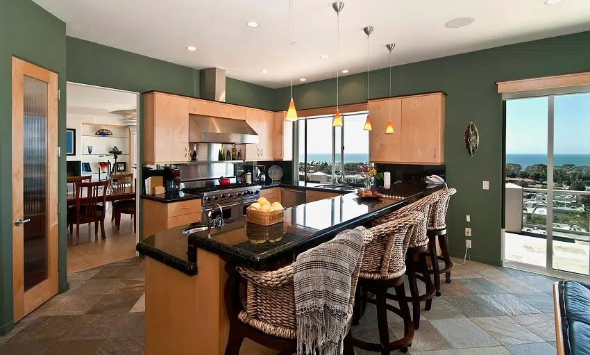 Apartment kitchen with slate tile floor green walls rattan armchairs black granite countertops pendant lights light wood cabinets and view of the dining area