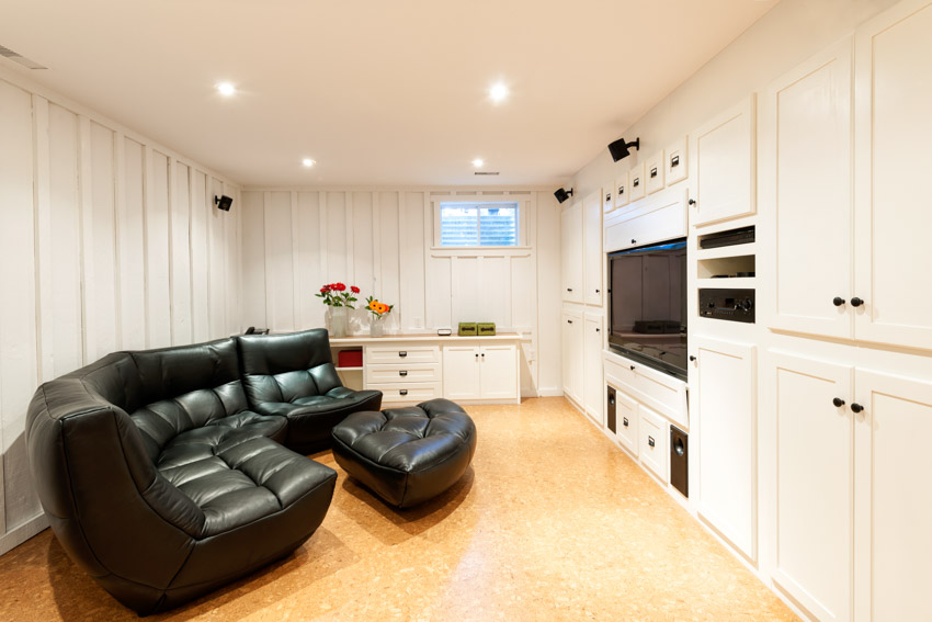 Basement living room with painted floor, recessed lights, black leather couch, and white cabinets