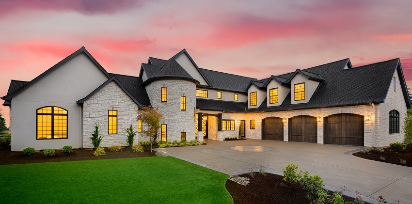 Black and white facade of stone mansion at sunset with manicured lawn concrete driveway wood garage doors and arch and columns at the entrance