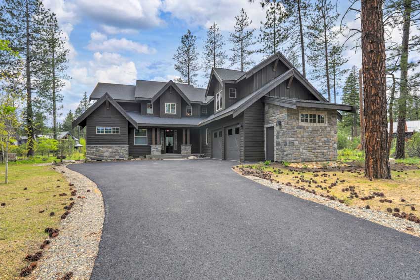 Black house with stone accent wall, pitched roof, and tar and chip driveway