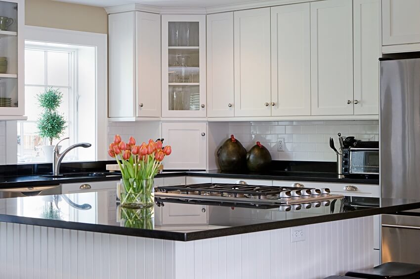 Bright kitchen with subway tile backsplash white cabinets black granite countertops and a beadboard island with a cooktop and flowers in a vase