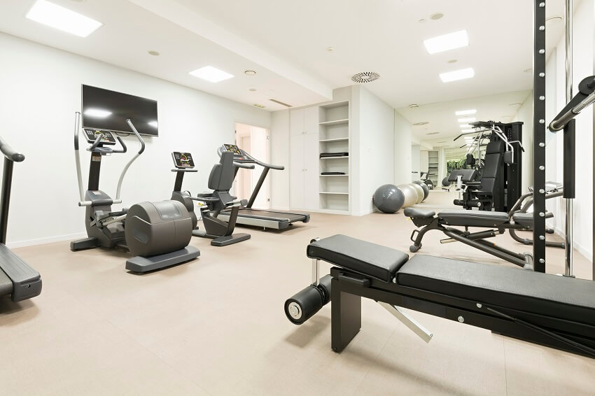 Bright white home gym with equipment carpet tile floor shelves and cabinets mirror and rectangular flush mount ceiling lighting
