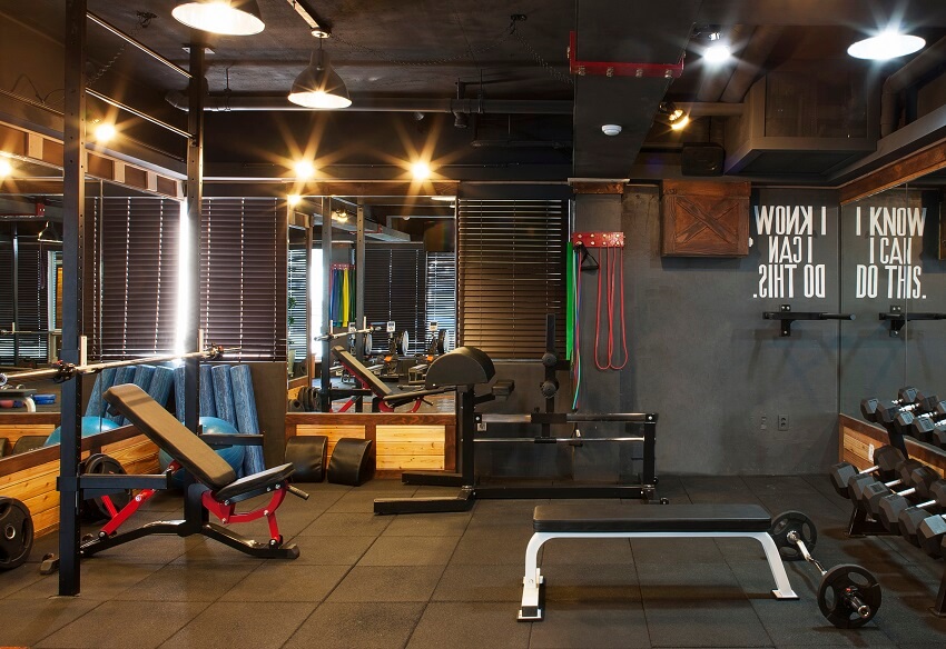 Garage gym with rubber mat tile floor dark grey walls mirrors overhead lighting gym equipment and windows with blinds