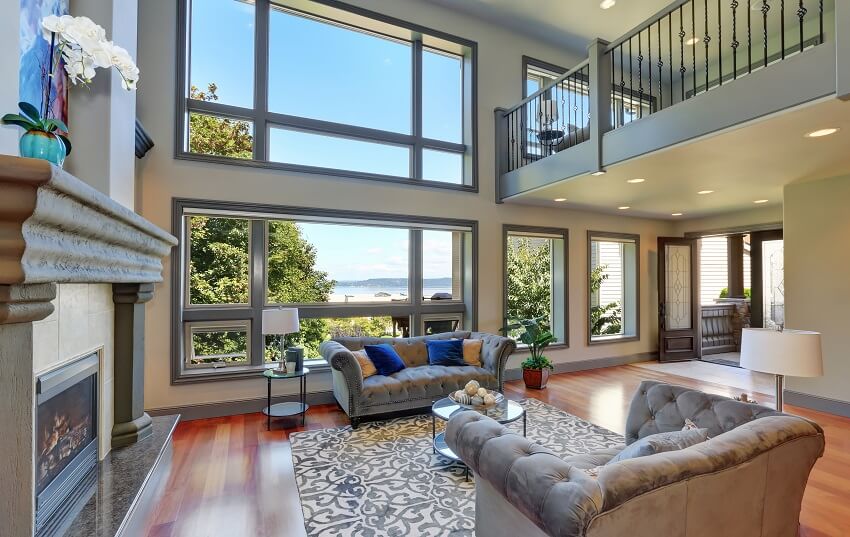 Grey interior of high vaulted ceiling living room in a mansion with hardwood floors fireplace a sofa with throw pillows and view of the beach through a large windows