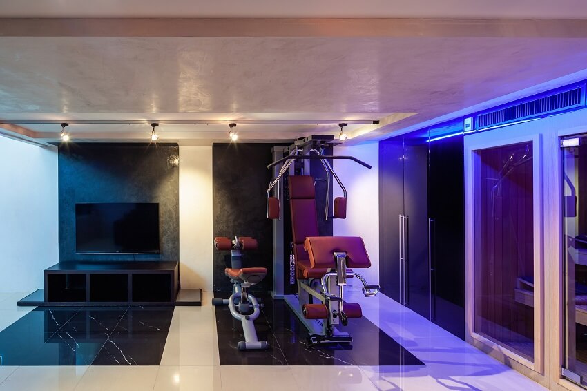 Home gym with marble tile floor track lighting black accent wall cubby hole storage with a wall mounted tv above gym equipment and sauna with glass doors