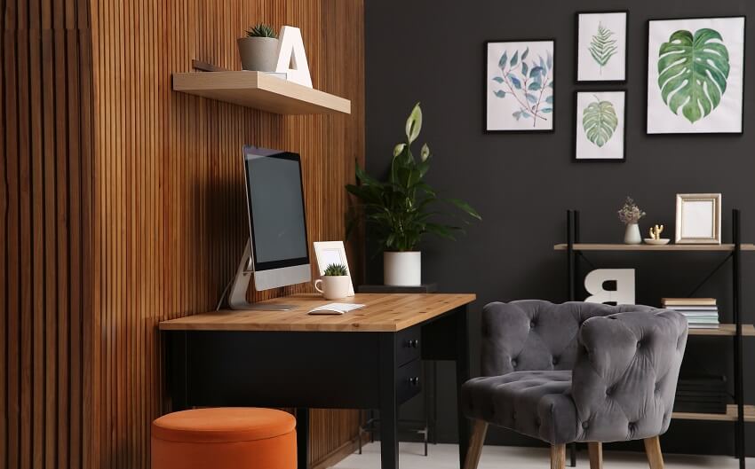 Home office design interior with computer on table near wooden wall grey armchair open and standing shelves orange ottoman and framed decors on grey wall