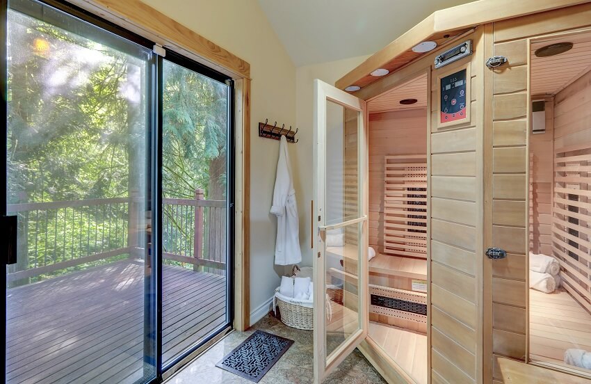 Home walk in large wooden sauna with open door and towels and view of a large deck with green trees through sliding doors