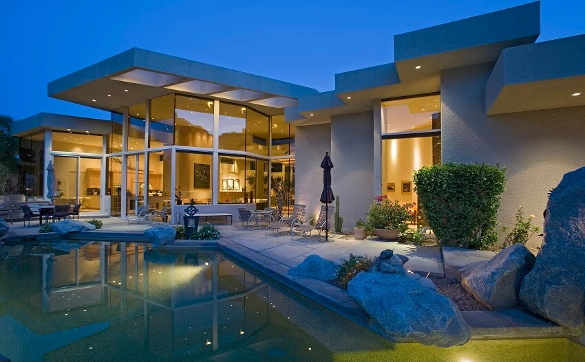 Illuminated glass and concrete house exterior with swimming pool in backyard pool side stones at dusk