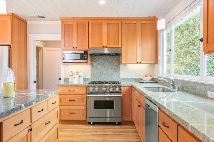 Kitchen with oak cabinets, black handles, wood flooring, windows, range hood, countertop, sink, faucet, and backsplash