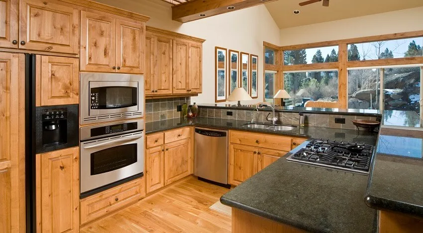 Kitchen With Wood Floors And Cabinets Grey Graninte Countertops With Cooktop Grey Stone Tile Backsplash And View Of The Living Room With Large Windows And Lamps Is .webp