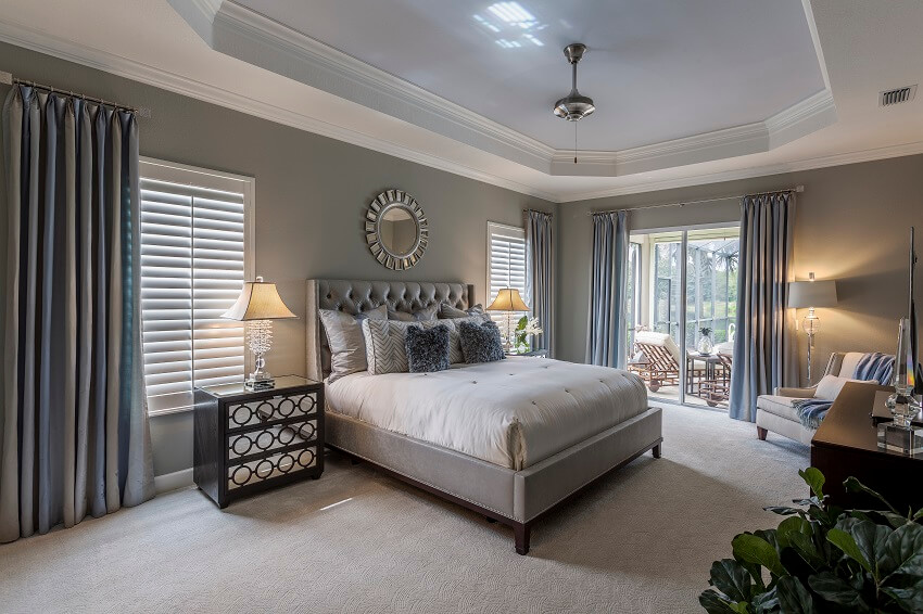 Large grey guest bedroom with carpeted floors lamps plant armchair wood furniture glass doors blue and grey curtains and a ceiling fan