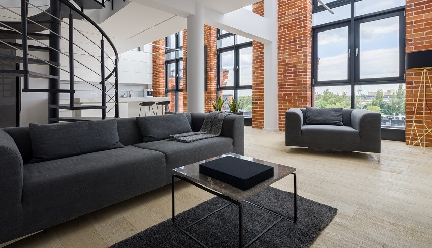 Living room in loft with big windows beams and columns brick walls black sofa coffee table on rug with pad metal stairs with railings hardwood floors and view of the kitchen