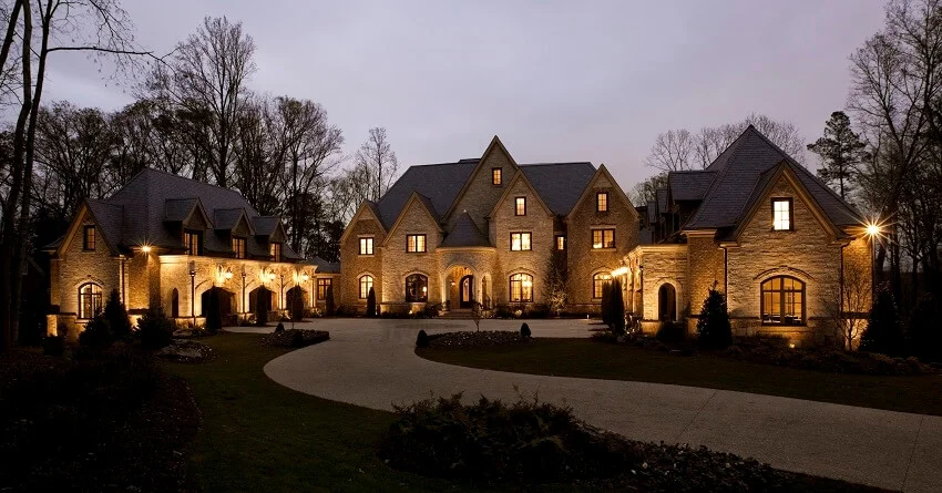 Long winding driveway leading up to the main entrance of a well lit stone mansion at dusk