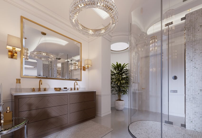 Modern bathroom with vanity and a mirror in a gold frame with sconces on the wall a low table with decor shower chandelier plant and brass sink handles