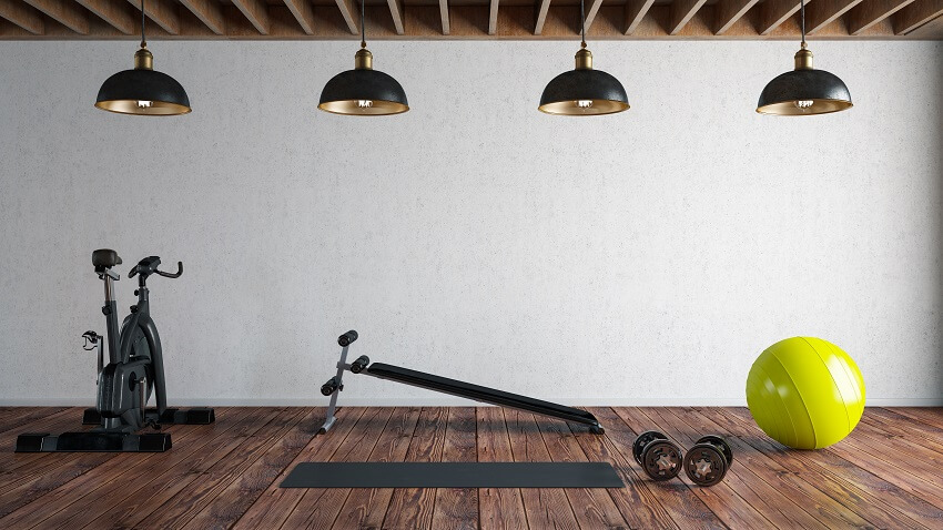 Modern home gym with hardwood floor four overhead lighting wood ceiling panels white empty wall and gym equipment