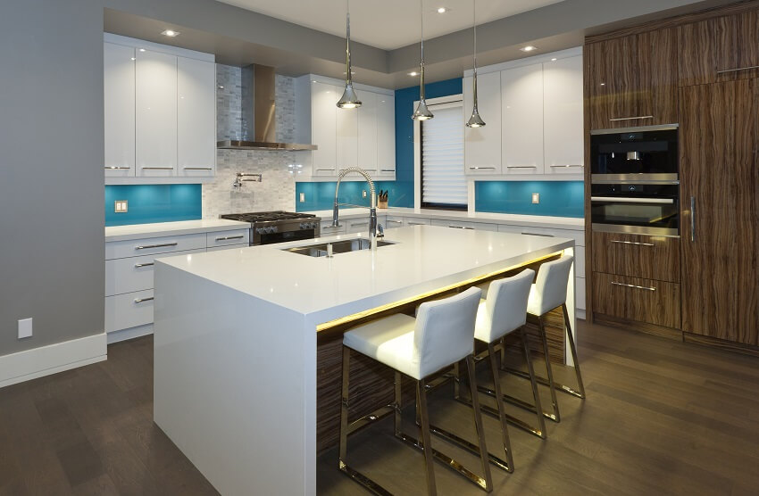 Modern kitchen with dark wood floor blue backsplash microwave drawer and pendant lights hanging above island white countertops sink and bar stools
