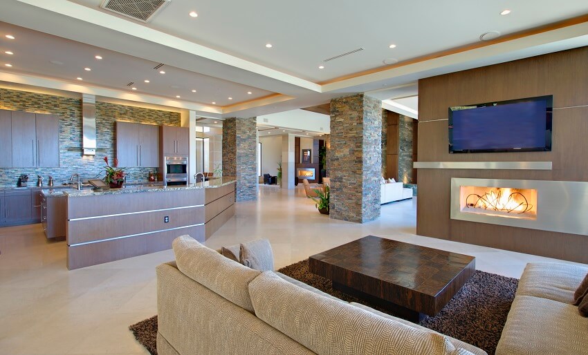 Modern living room with open kitchen stone columns and backsplash tile floors fireplace wood kitchen cabinets sofa coffee table and recessed lighting