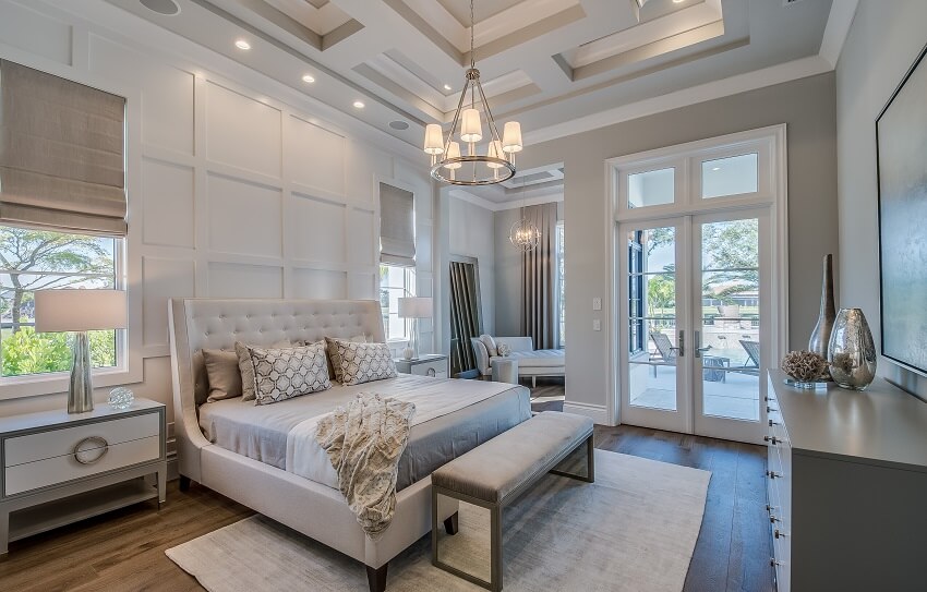 White guest bedroom with high coffered ceiling wainscoting wall hardwood floors lighting fixtures bedroom bench with mirror a couch and french doors