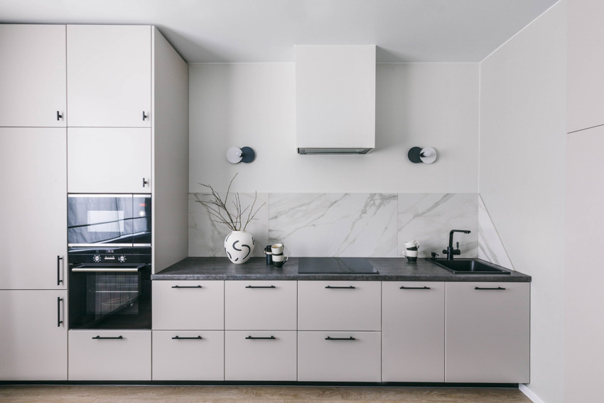 White flat panel kitchencabinets, veined stone backsplash, oven, and black countertop