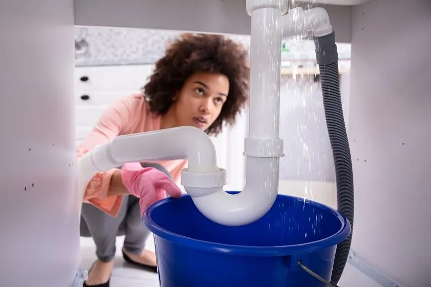 Woman looking at water leaking from sink pipe to a blue bucket under countertop 
