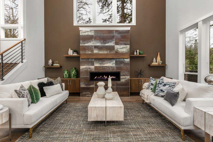 Beautiful living room with brown accent wall, fireplace, shelves, white couches, rug, staircase, and glass windows