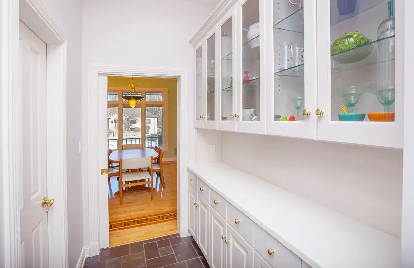 A minimalist built in pantry with white cabinet storage, floor tiles and view from dining area