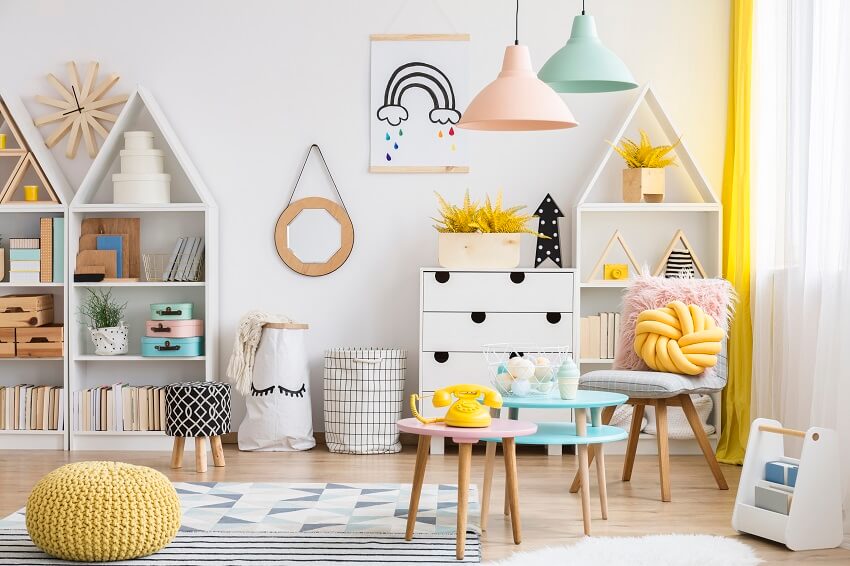 Pastel playroom with yellow pouf near pink table pendant lights poster on white wall and book shelves