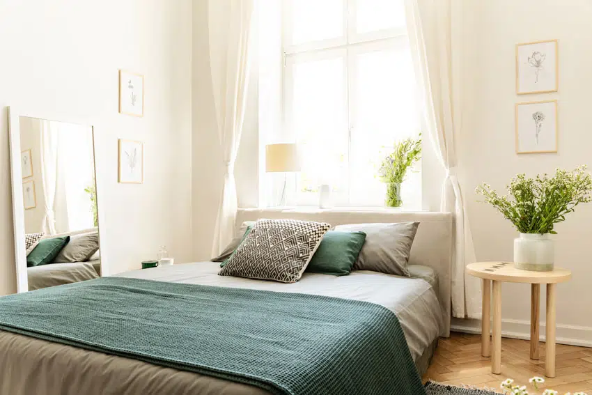 Small guest bedroom with green bedsheets, pillows, nightstand, mirror, and window curtain