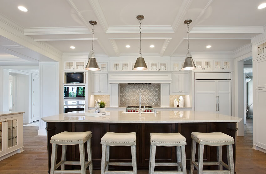 White kitchen with barstools in brown island, coffered ceiling, metallic backsplash behind stove