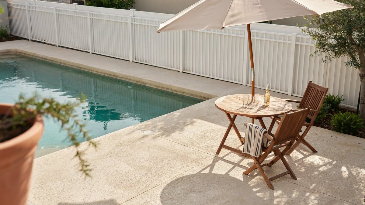 Peaceful poolside scene with a fence, umbrella, table and chairs