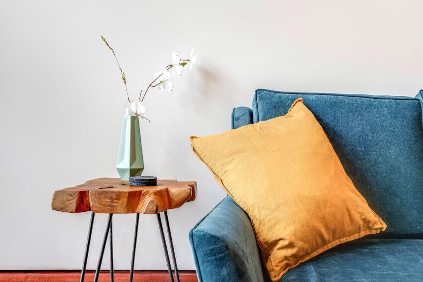 A wood plank table with a vase and flowers on top and a turquoise couch with a yellow pillow