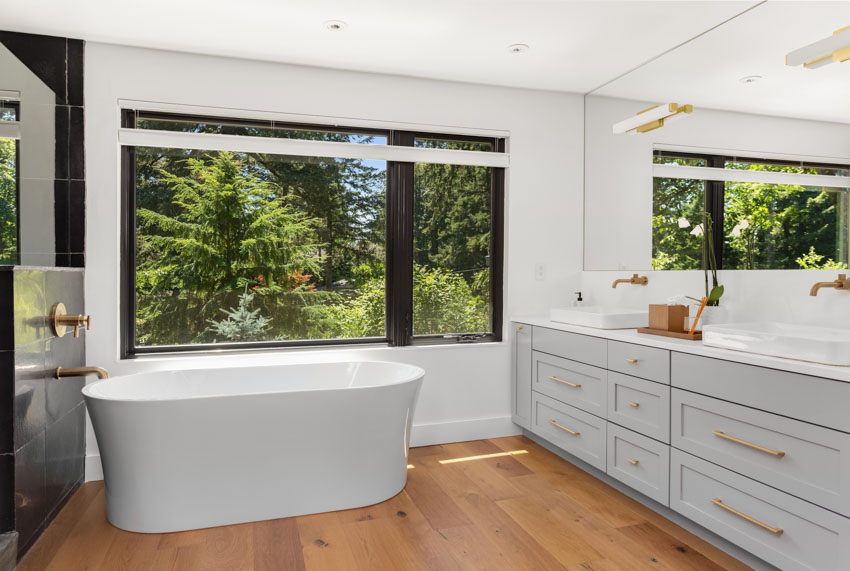 Bathroom with acrylic tub, drawers, mirror, windows, and wood flooring