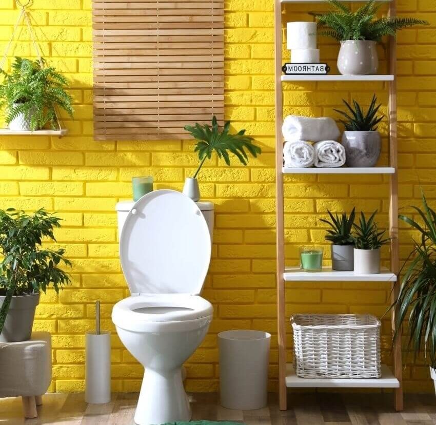Bathroom with toilet bowl decors on toilet tank and green plants near yellow brick wall