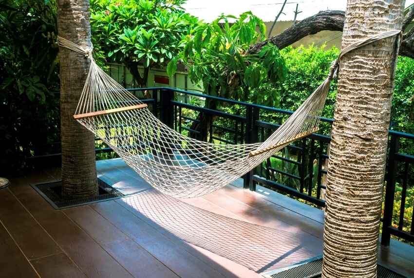 Cotton rope hammock hanging in between two palm trees at the wooden terrace near outdoor garden