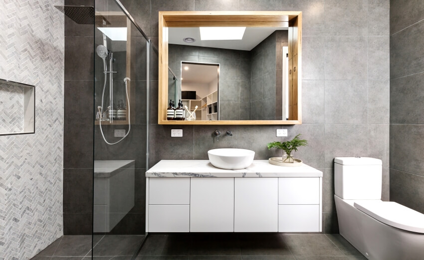 Grey bathroom with herringbone shower tiling, wood framed mirror, and a floating marble countertop