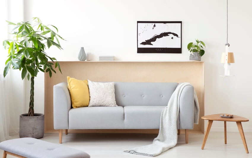 Grey sofa with cushions and blanket in white living room with plants and wooden triangle end table