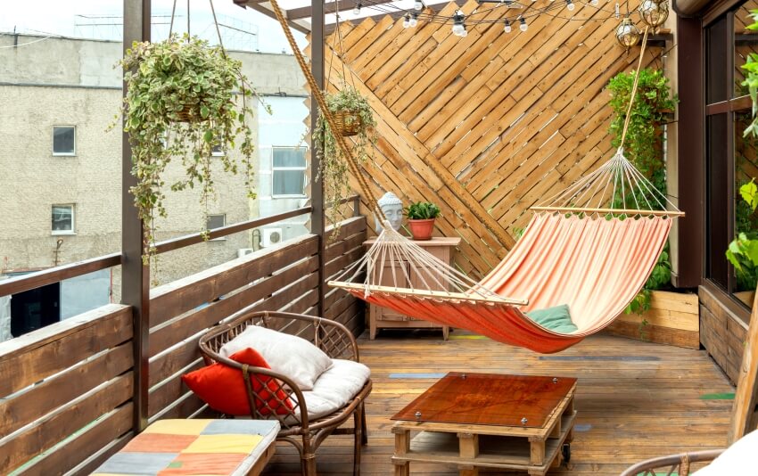 Hammock hangs on wooden terrace with table, flower pots, chairs, and old buildings in the background