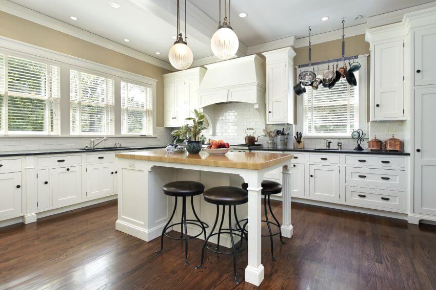 Kitchen with wood countertop island, white cabinets with black hardware, and windows with blinds