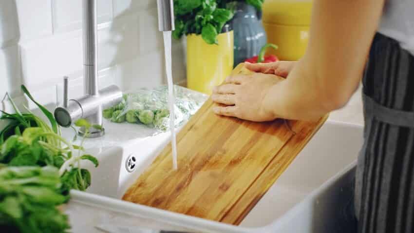 A person washing an acacia cutting board with a cleaning liquid under tap water