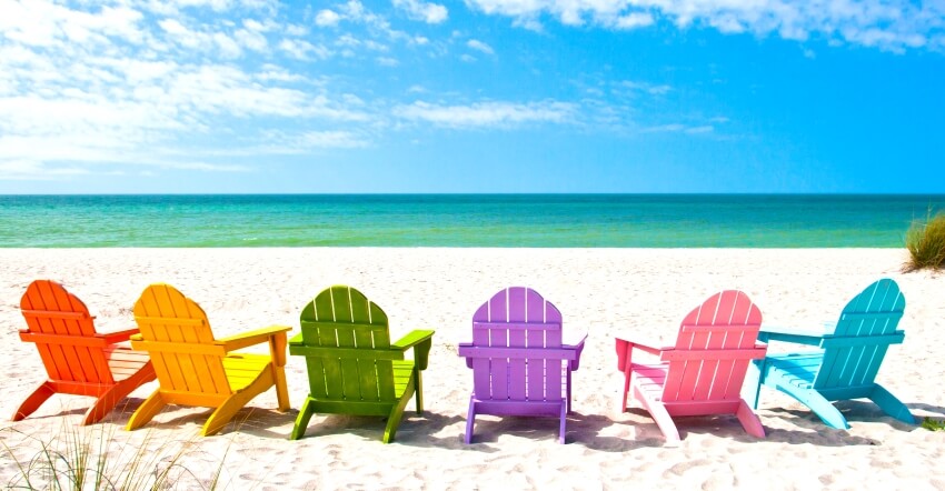 A rainbow colored row of adirondack chairs on a sunny vacation beach